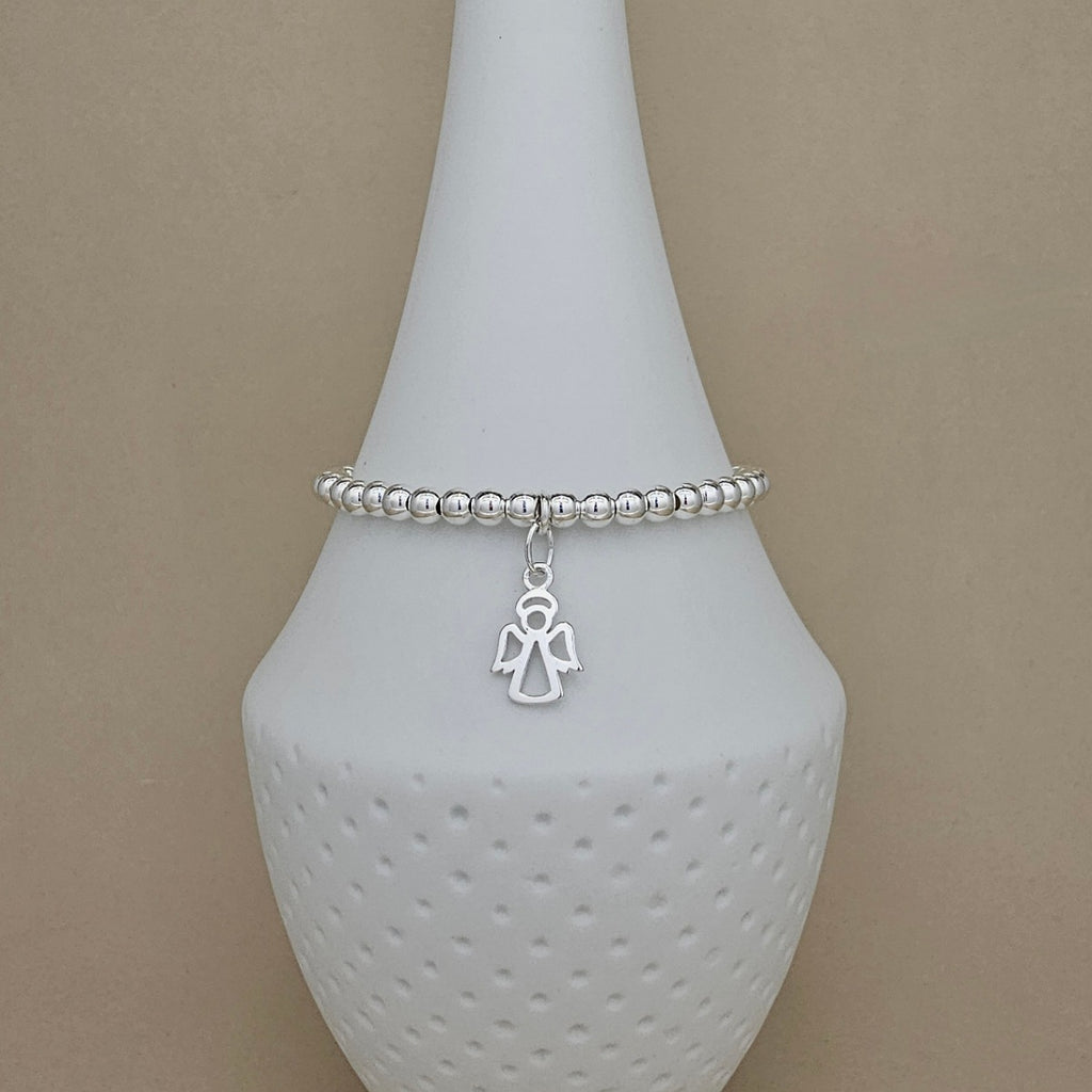 A silver beaded bracelet with a small angel charm, displayed on a white ceramic jewelry stand against a neutral beige background.