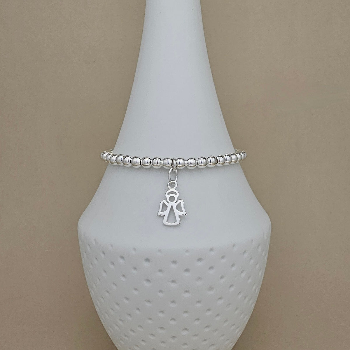 A silver beaded bracelet with a small angel charm, displayed on a white ceramic jewelry stand against a neutral beige background.