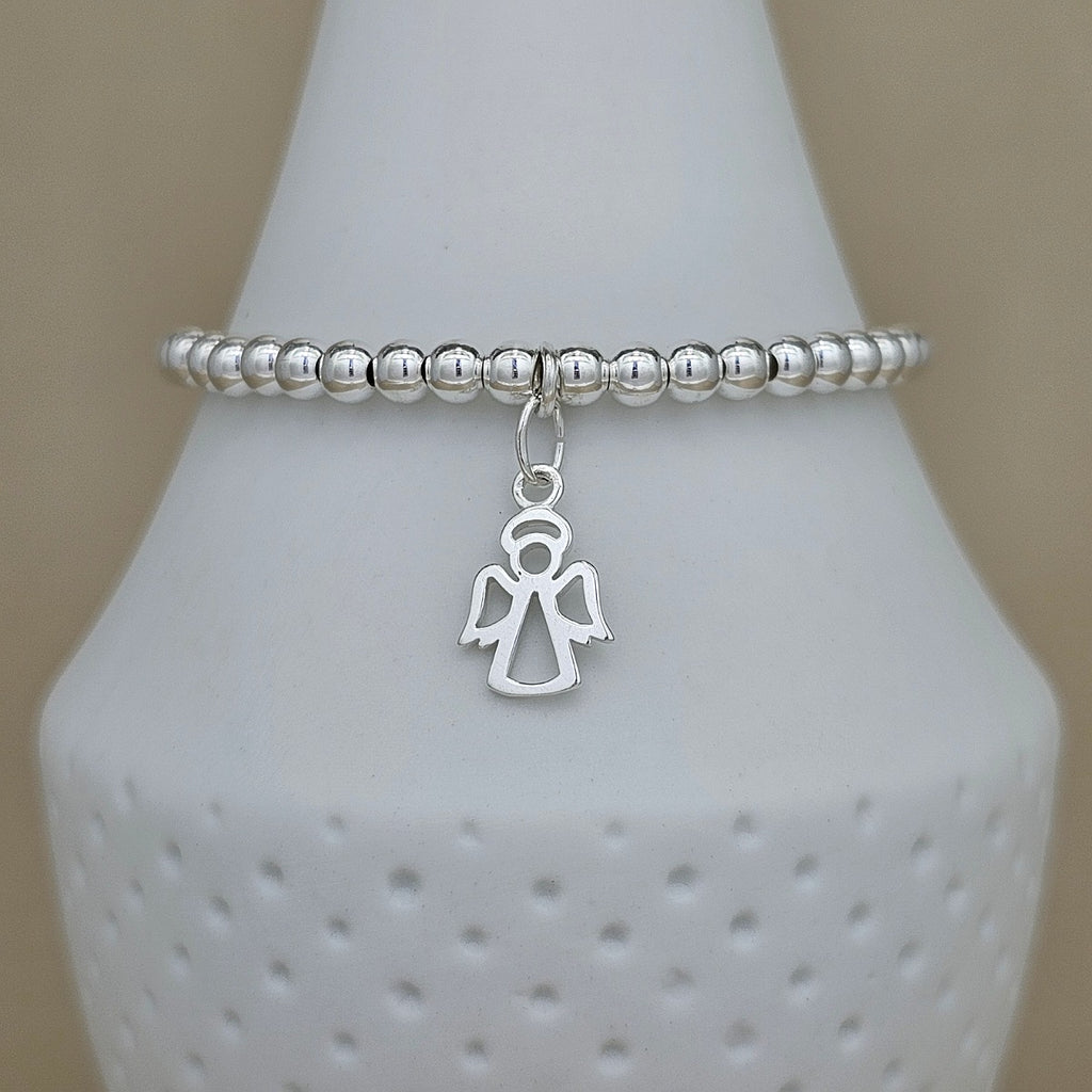 A silver beaded stretch bracelet with a dangling angel charm, displayed on a white ceramic cone stand against a neutral beige background.
