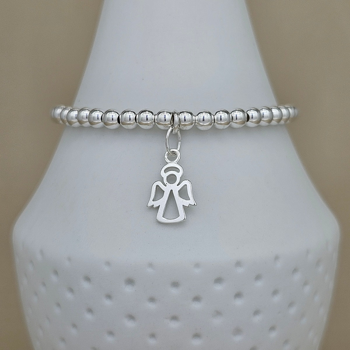 A silver beaded stretch bracelet with a dangling angel charm, displayed on a white ceramic cone stand against a neutral beige background.