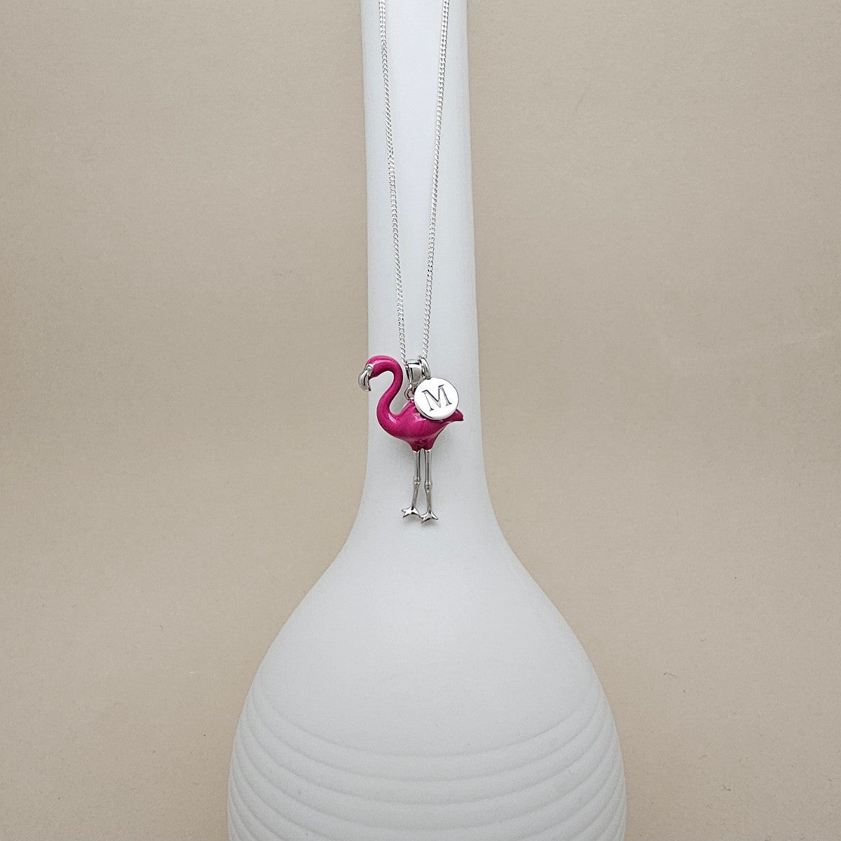 A silver necklace with a pink flamingo pendant holding a small silver heart engraved with the letter 'M', displayed on a white ribbed vase against a beige background.