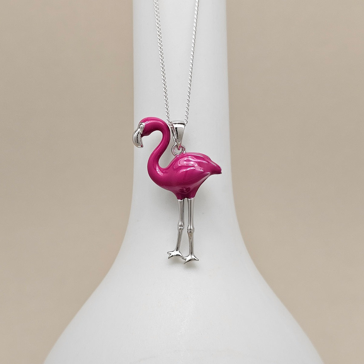 A silver flamingo pendant necklace with a vibrant magenta-pink body, displayed on a white ceramic stand against a warm beige background.