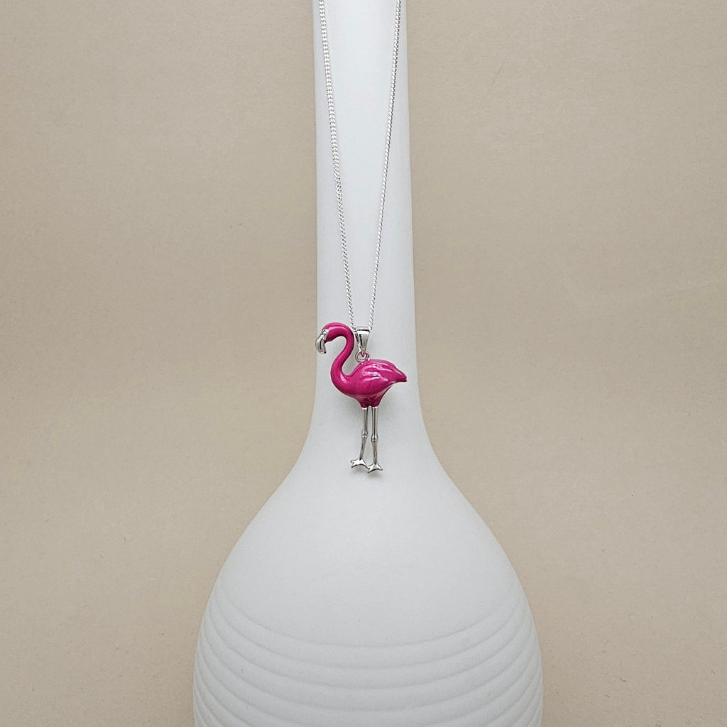 A silver-toned necklace with a bright pink flamingo pendant, displayed on a white ribbed ceramic vase against a neutral beige background.