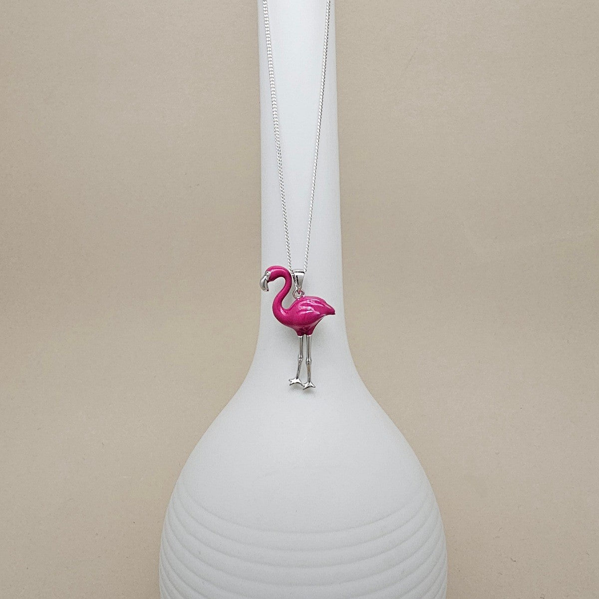 A silver-toned necklace with a bright pink flamingo pendant, displayed on a white ribbed ceramic vase against a neutral beige background.