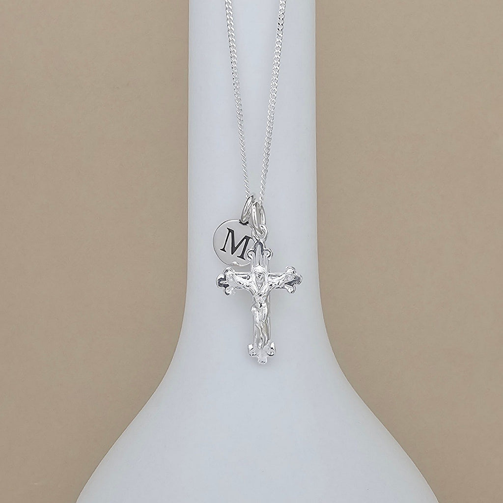 A silver cross necklace with a crucifix pendant and a round initial charm engraved with 'M', displayed on a white neck mannequin against a beige background.