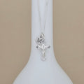 A silver cross necklace with a crucifix pendant and a round initial charm engraved with 'M', displayed on a white neck mannequin against a beige background.