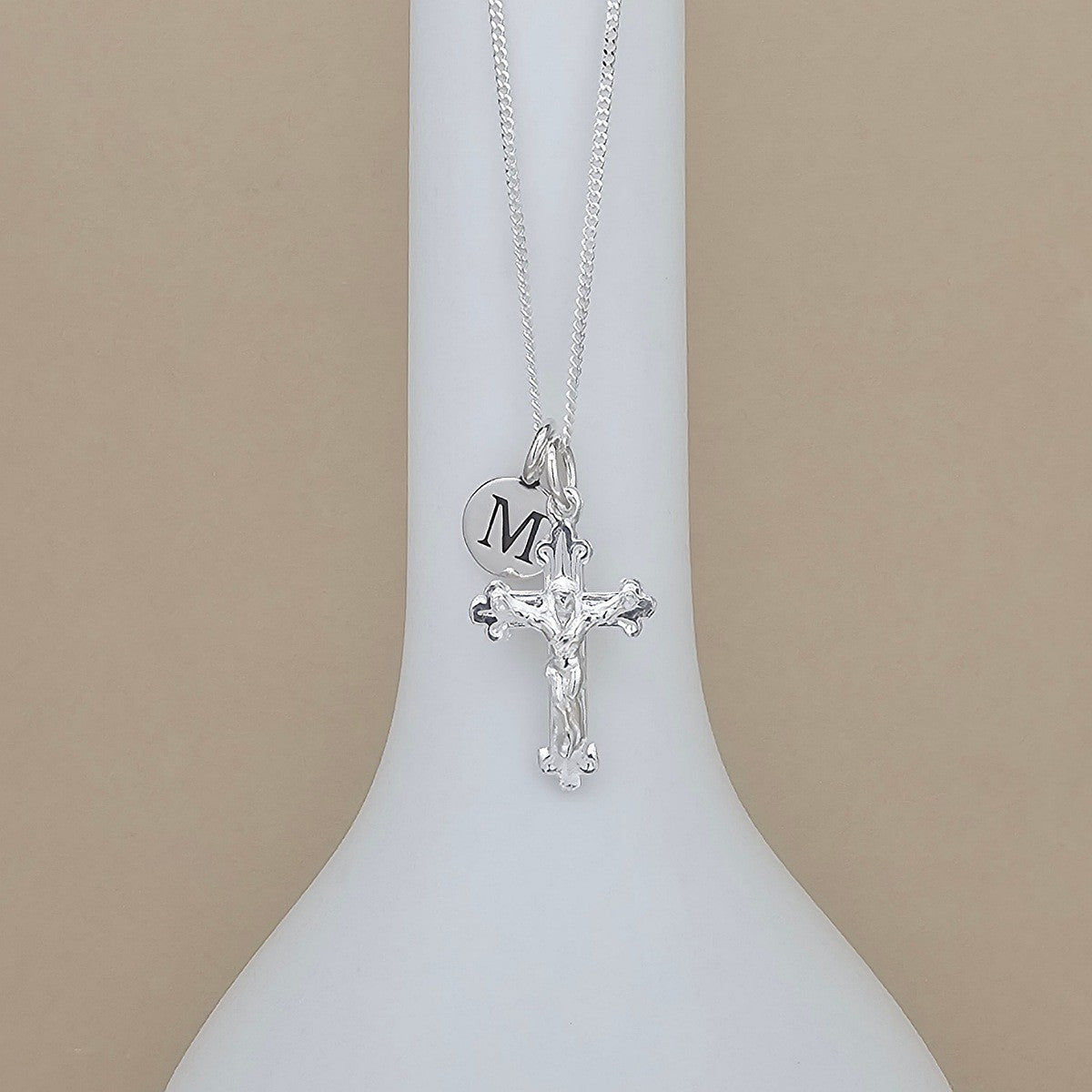 A silver cross necklace with a crucifix pendant and a round initial charm engraved with 'M', displayed on a white neck mannequin against a beige background.