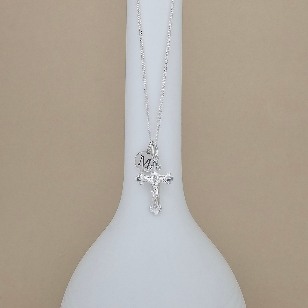 A silver crucifix pendant necklace with a small heart charm engraved with the initial 'M', displayed on a white vase against a beige background.