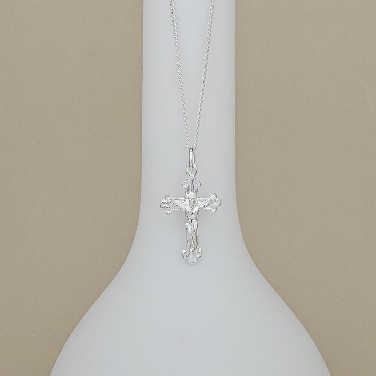 A silver crucifix pendant necklace displayed on a white neck mannequin against a solid beige background.