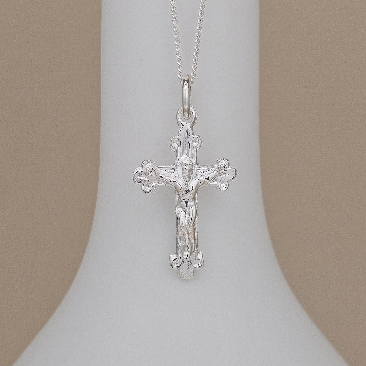 A silver crucifix pendant necklace displayed on a white cone-shaped jewelry bust against a soft beige background.