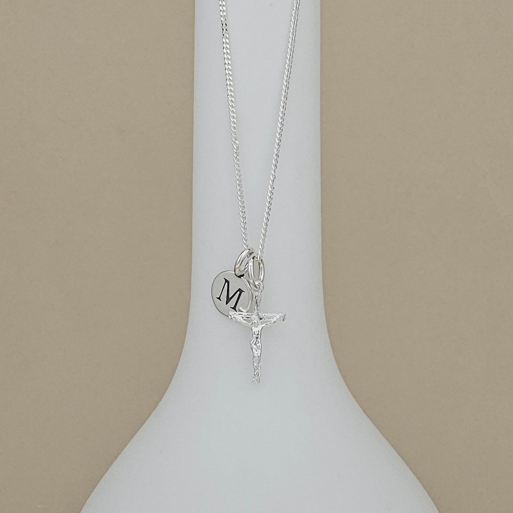 A silver necklace with a crucifix pendant and a heart-shaped initial 'M' charm, displayed on a white ceramic stand against a beige background.