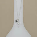 A silver necklace with a crucifix pendant and a heart-shaped initial 'M' charm, displayed on a white ceramic stand against a beige background.