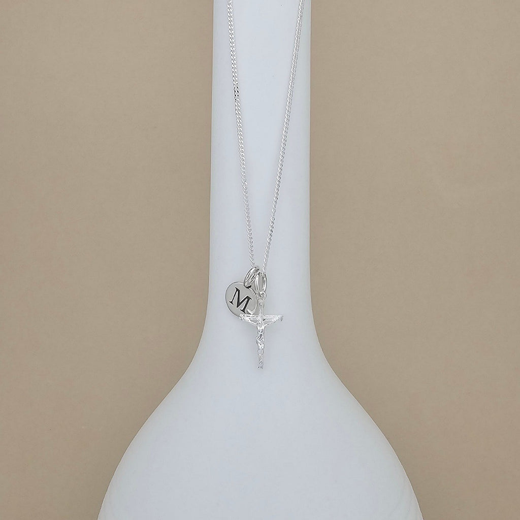 A silver necklace with a round pendant engraved with the letter 'M' and a small crucifix charm, displayed on a white ceramic vase against a beige background.
