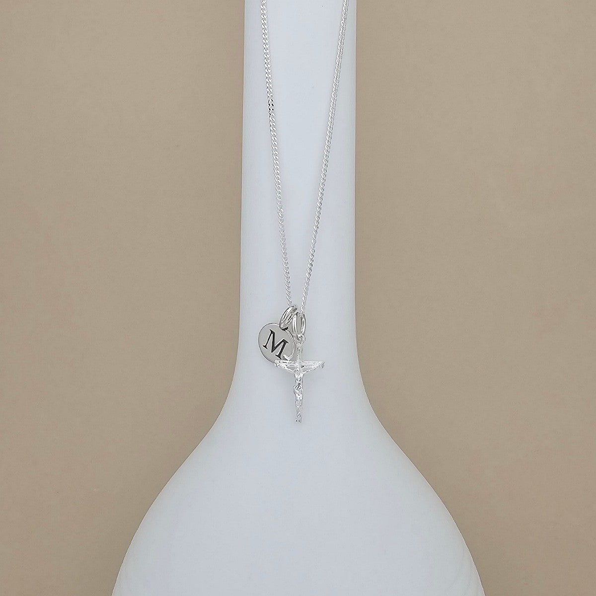 A silver necklace with a round pendant engraved with the letter 'M' and a small crucifix charm, displayed on a white ceramic vase against a beige background.