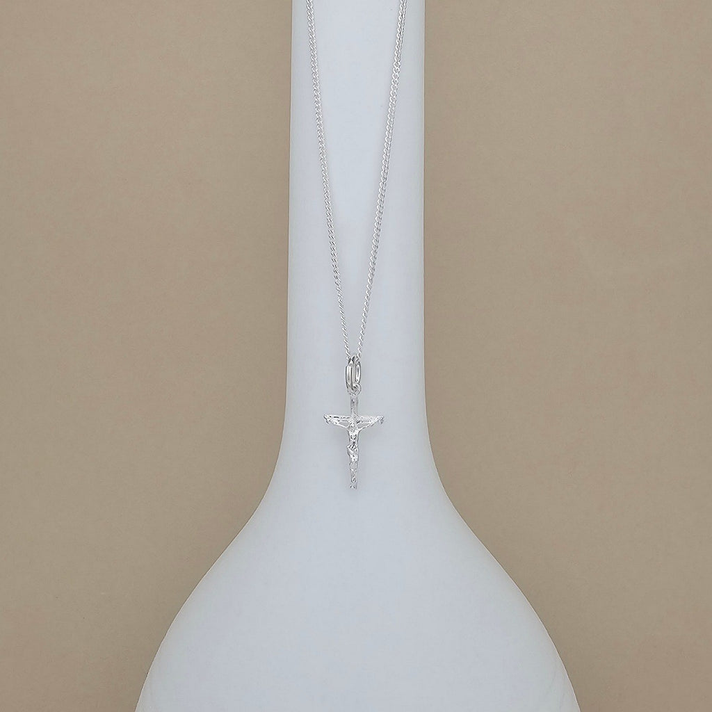 A silver crucifix pendant necklace on a delicate silver chain, displayed on a white ceramic vase against a solid beige background.