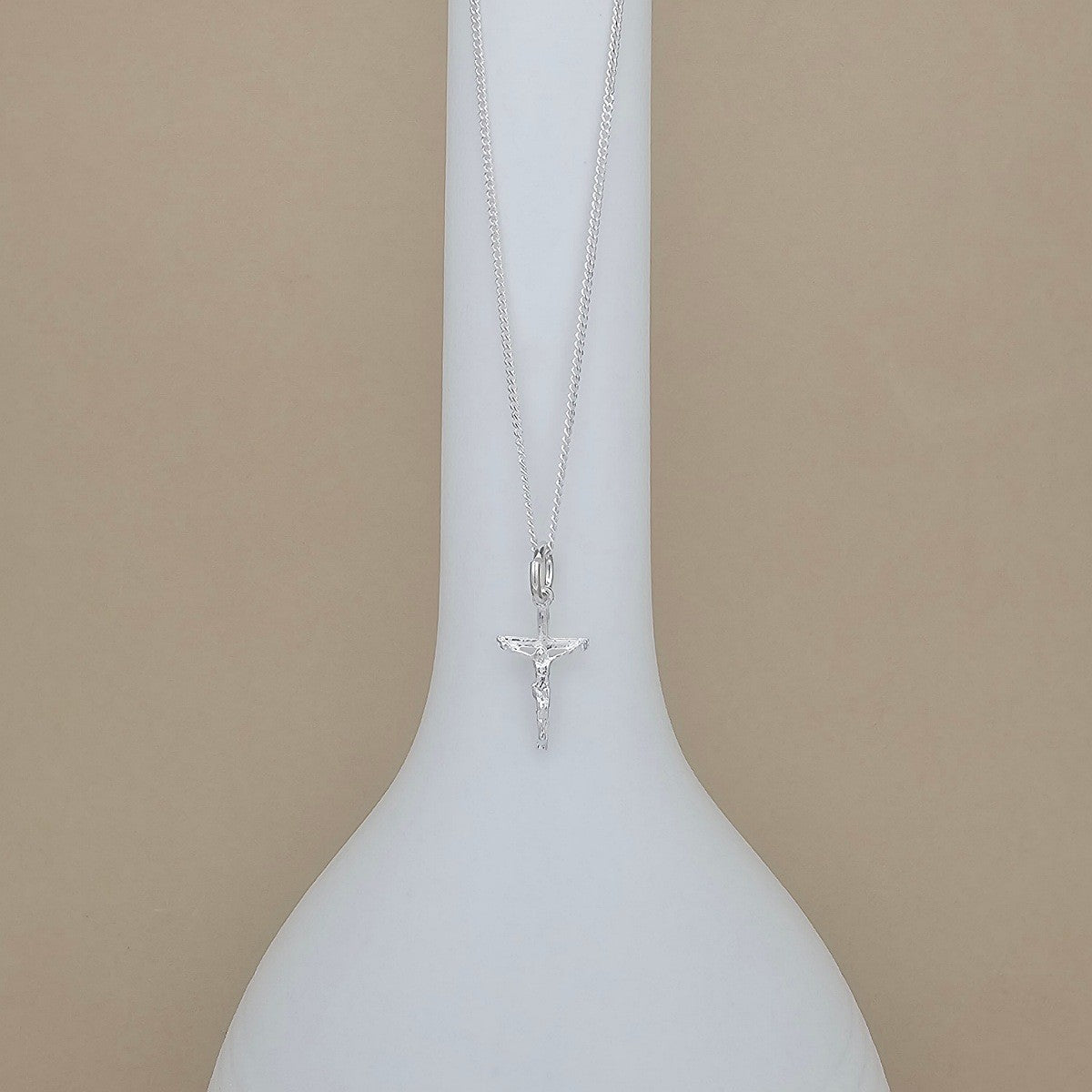A silver crucifix pendant necklace on a delicate silver chain, displayed on a white ceramic vase against a solid beige background.