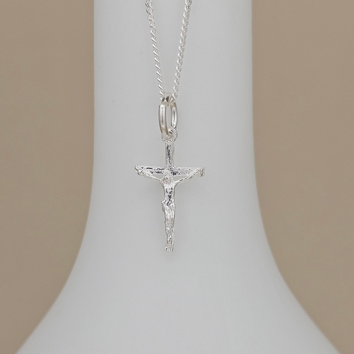 A silver crucifix pendant necklace displayed on a white jewelry bust against a soft beige background.