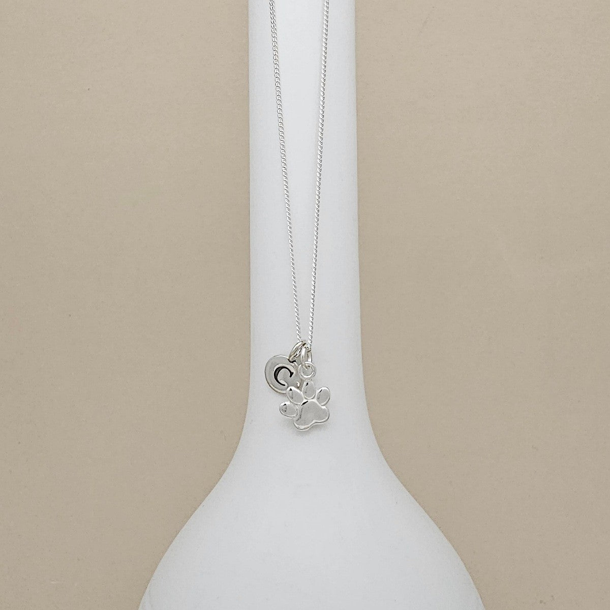 A silver necklace with a heart and paw print pendant, displayed on a white ceramic stand against a beige background.