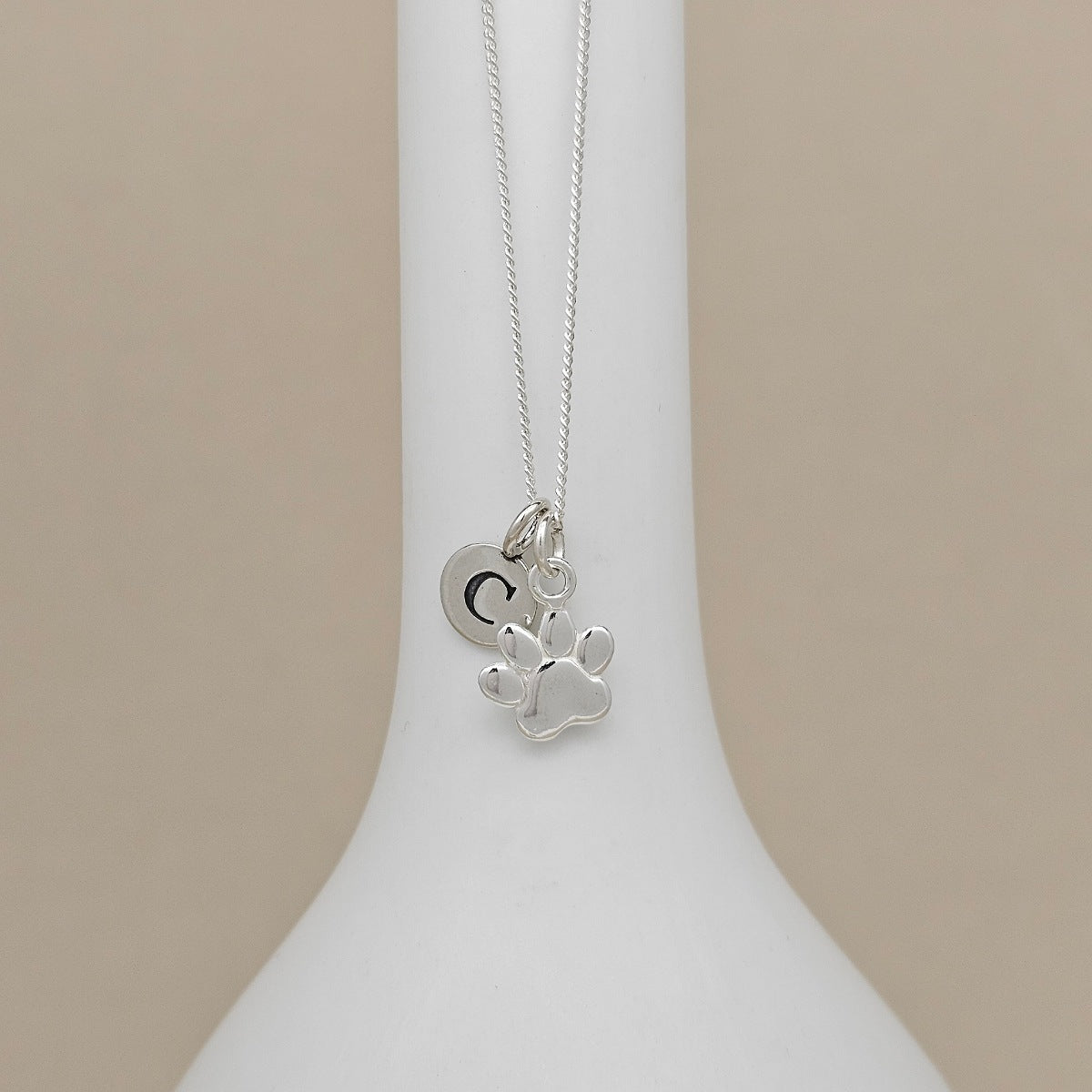A silver necklace with a paw print and crescent moon pendant, displayed on a white ceramic stand against a warm beige background.