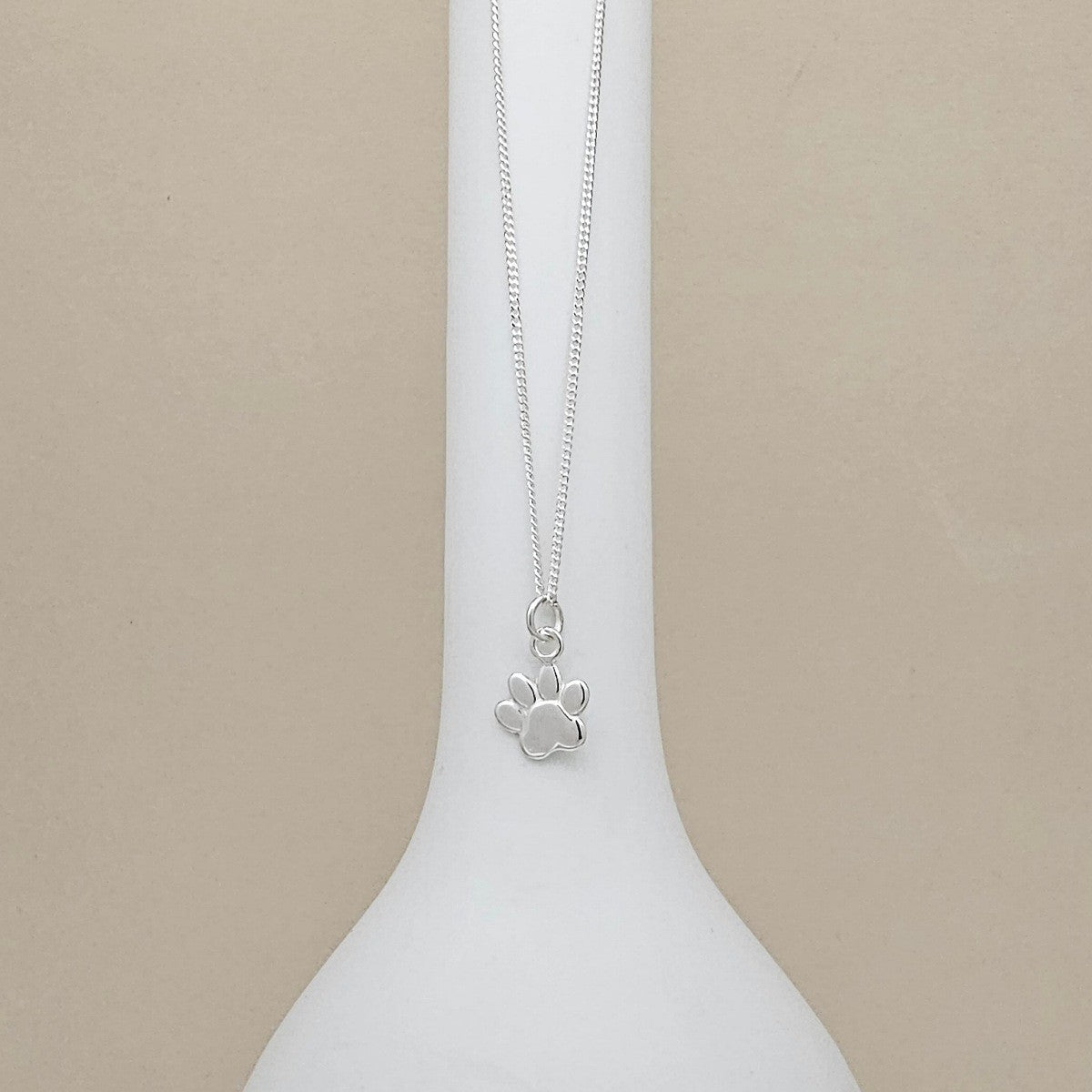 A silver chain necklace with a small paw print pendant, displayed on a white ceramic vase against a solid beige background.