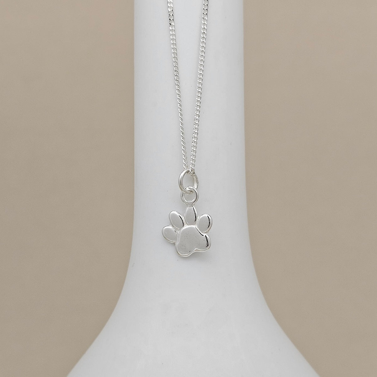 A silver paw print pendant necklace displayed on a white neck stand against a neutral beige background.