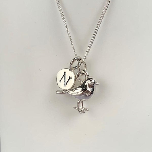 A silver necklace featuring a bird charm and a round initial pendant engraved with the letter 'N', displayed against a plain light background.