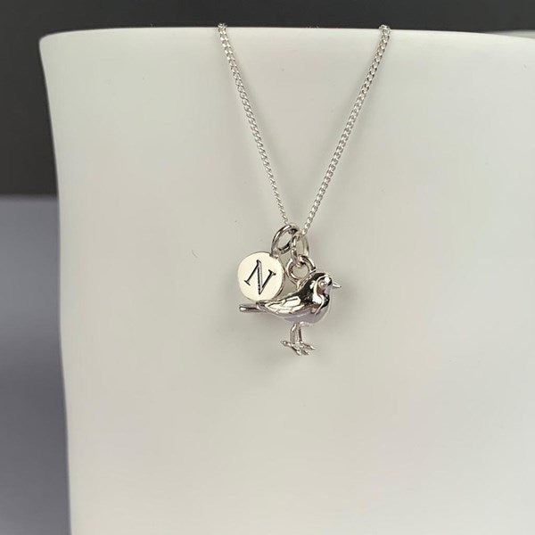 A silver necklace with a bird charm and a round engraved disc charm featuring the letter 'N', displayed on a white jewelry bust.