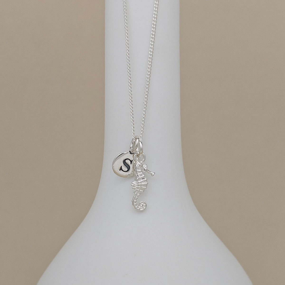 A silver necklace with a seahorse and initial 'S' charm, displayed on a white ceramic stand against a beige background.