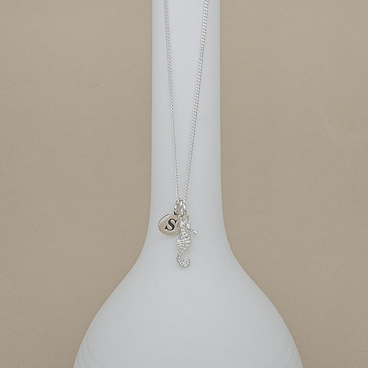 A silver necklace with a seahorse and initial 'S' charm, displayed on a white ceramic vase against a beige background.