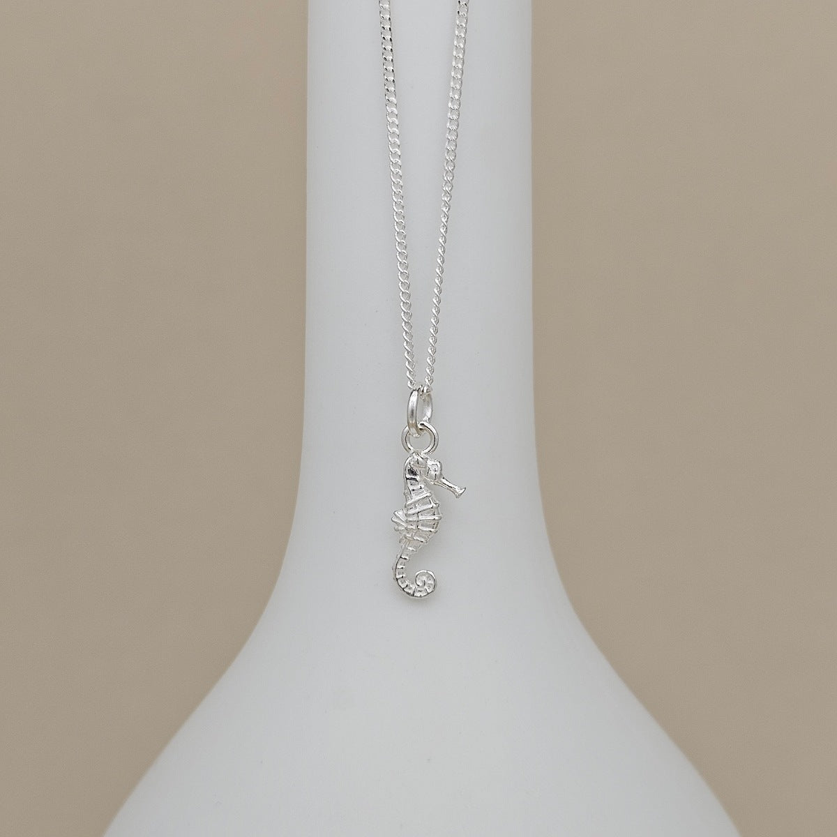 A silver seahorse pendant necklace displayed on a white ceramic vase against a solid beige background.