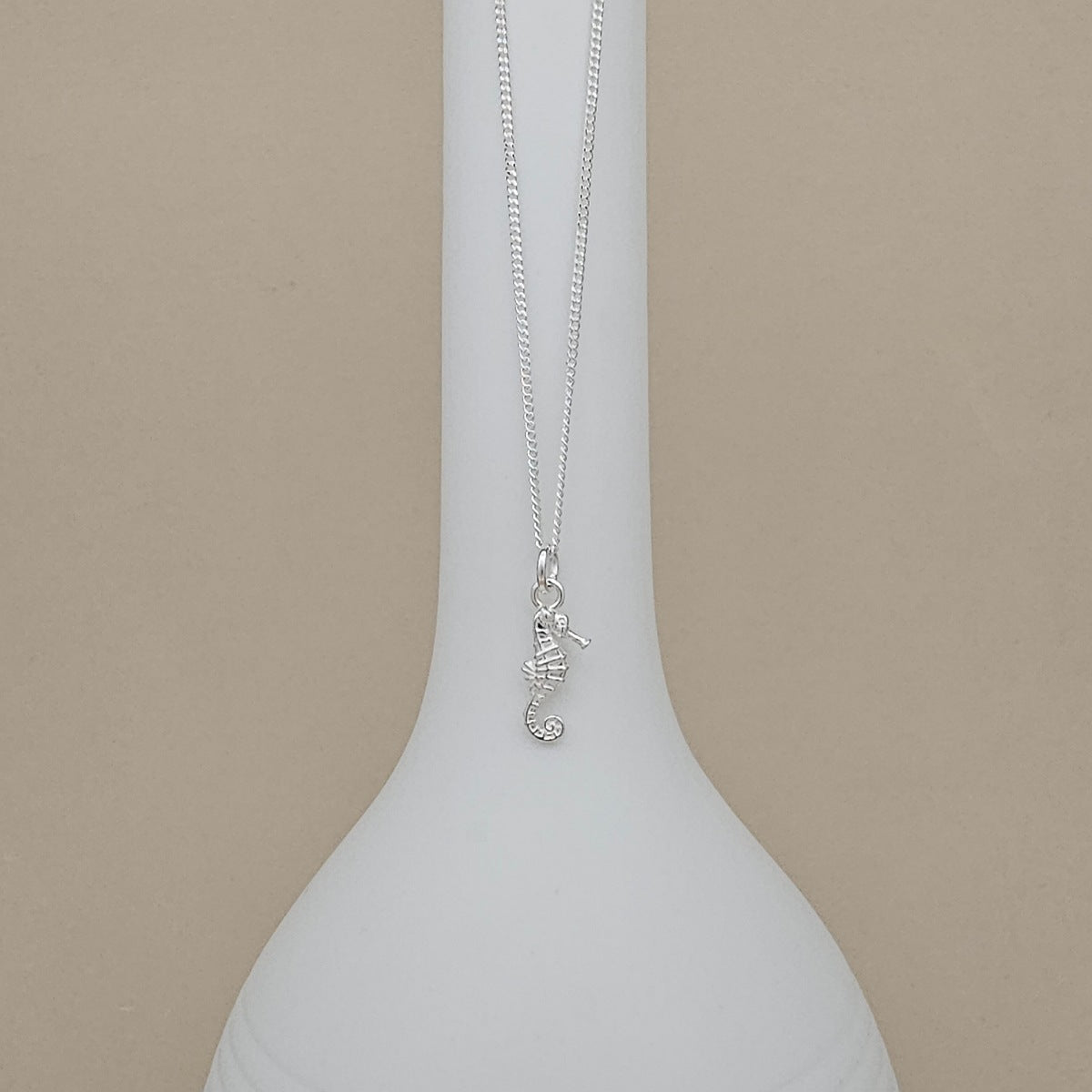 A silver seahorse pendant necklace displayed on a white ceramic vase against a solid beige background.