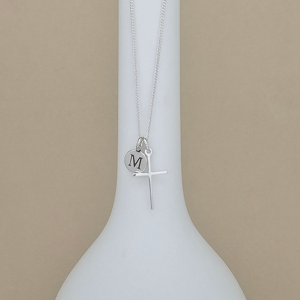A silver necklace with a cross pendant and a small round initial 'M' disc charm, displayed on a white bottle-shaped stand against a beige background.