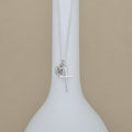 A silver necklace with a cross pendant and a small round initial 'M' disc charm, displayed on a white bottle-shaped stand against a beige background.