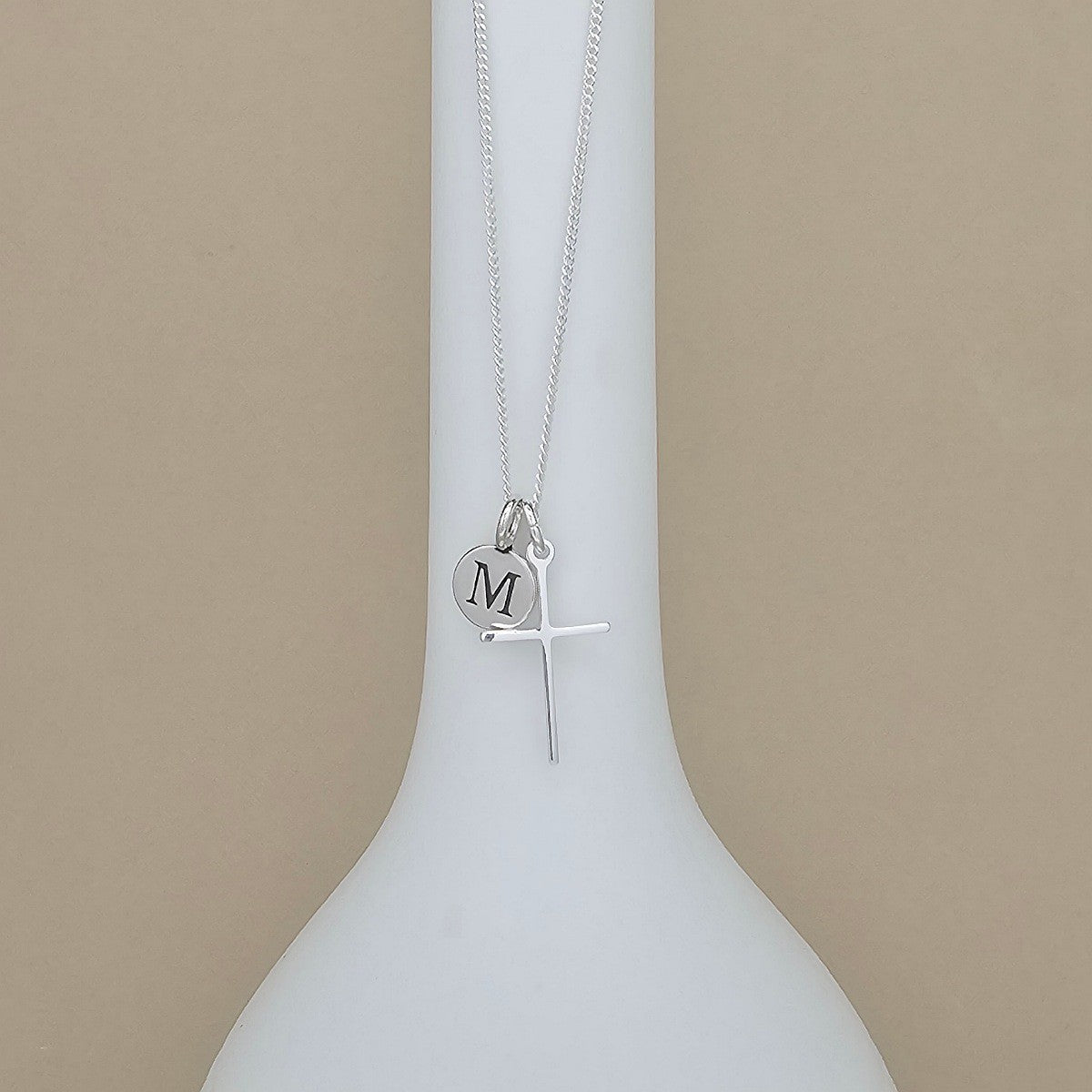 A silver necklace with a cross pendant and a small round initial 'M' disc charm, displayed on a white bottle-shaped stand against a beige background.