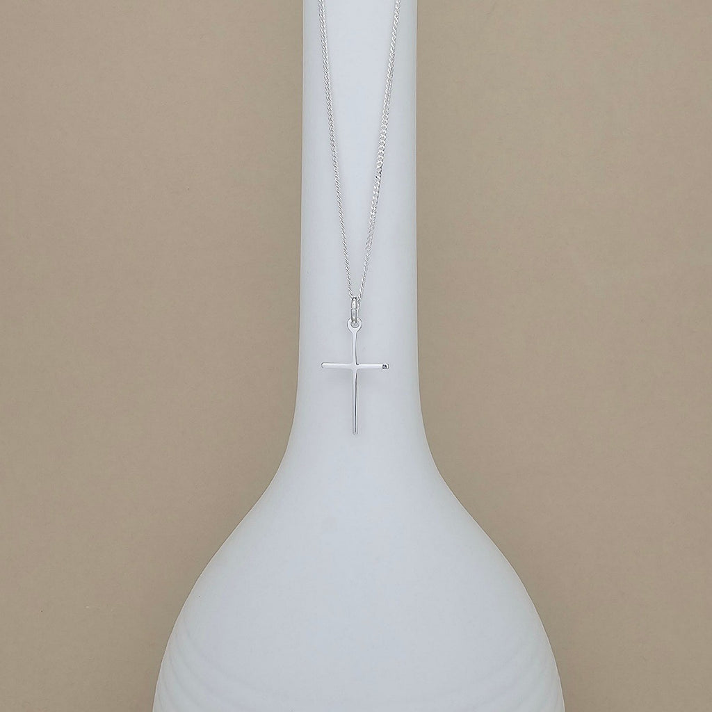 A silver cross pendant necklace hanging from a delicate chain, displayed against a white ceramic vase on a warm beige background.