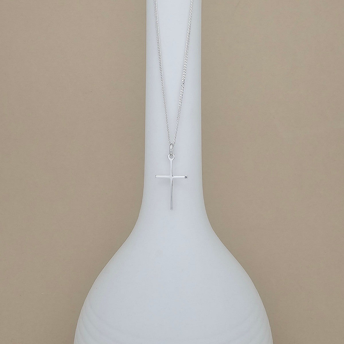 A silver cross pendant necklace hanging from a delicate chain, displayed against a white ceramic vase on a warm beige background.