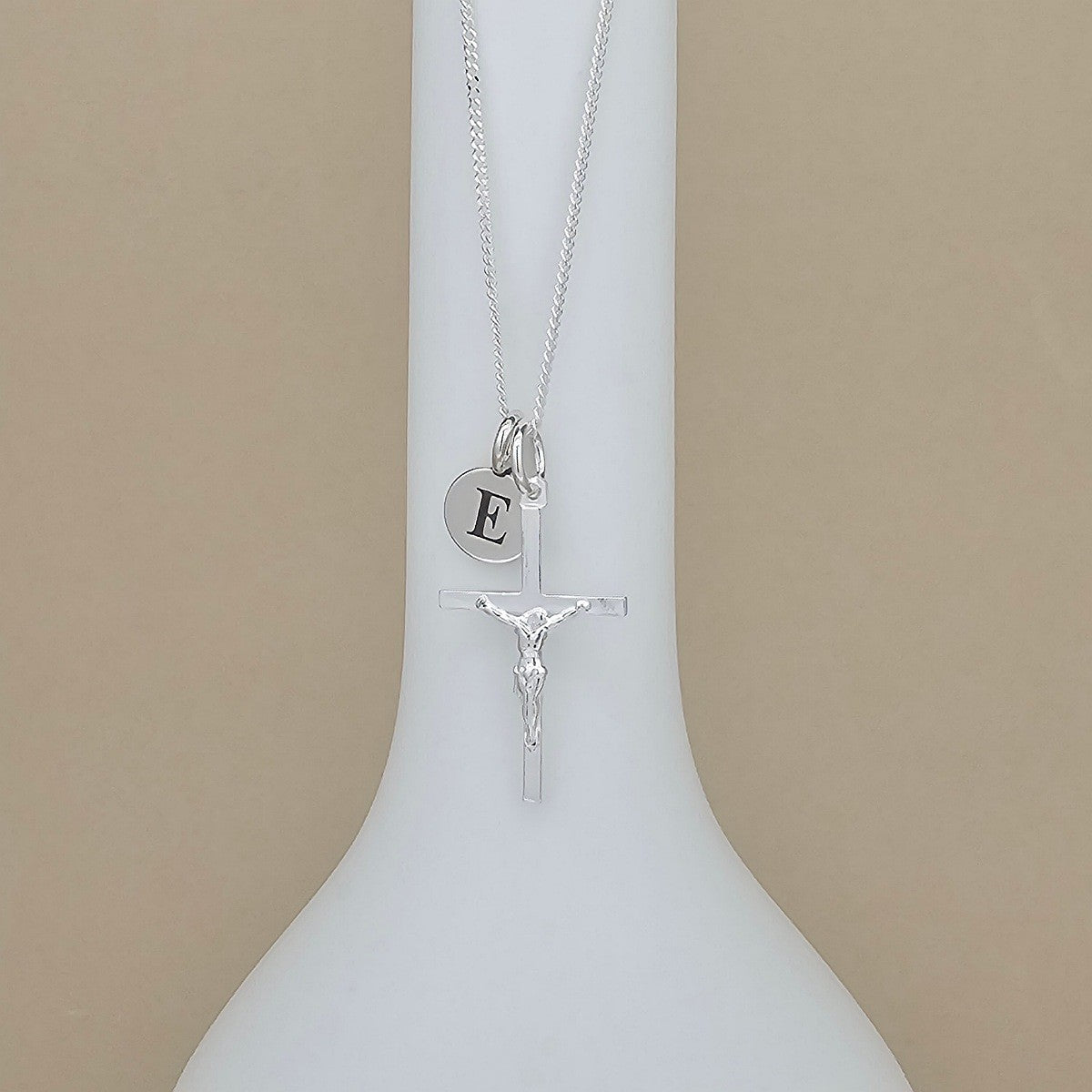 A silver necklace displayed on a white vase, featuring a crucifix pendant, a round initial 'E' charm, and a delicate chain against a beige background.