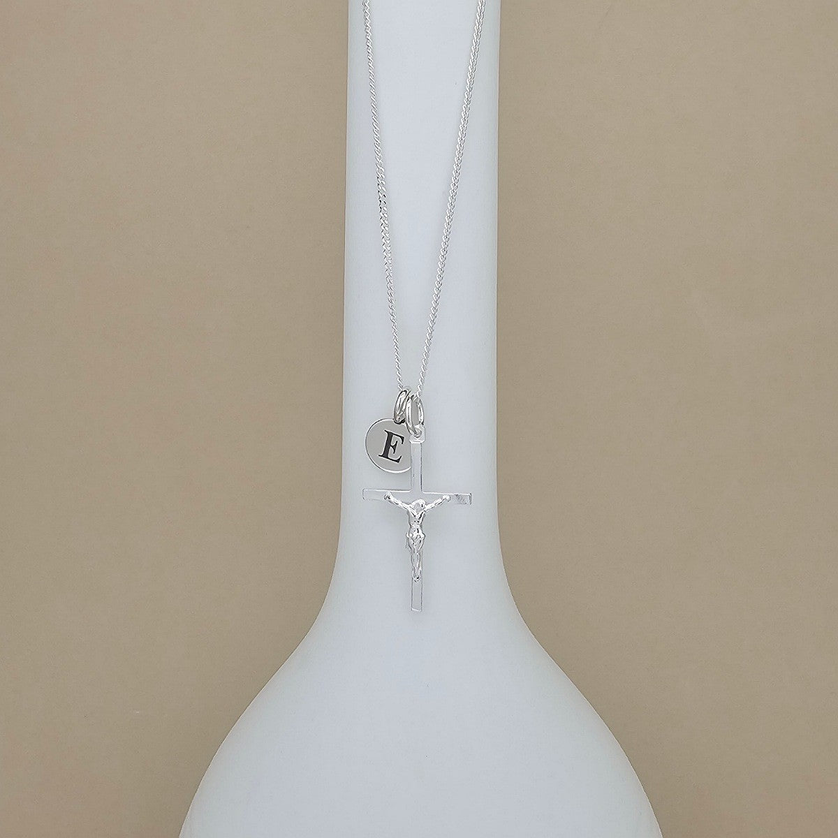 A silver necklace with a crucifix pendant and a small round initial pendant engraved with the letter 'E', displayed on a white vase against a beige background.