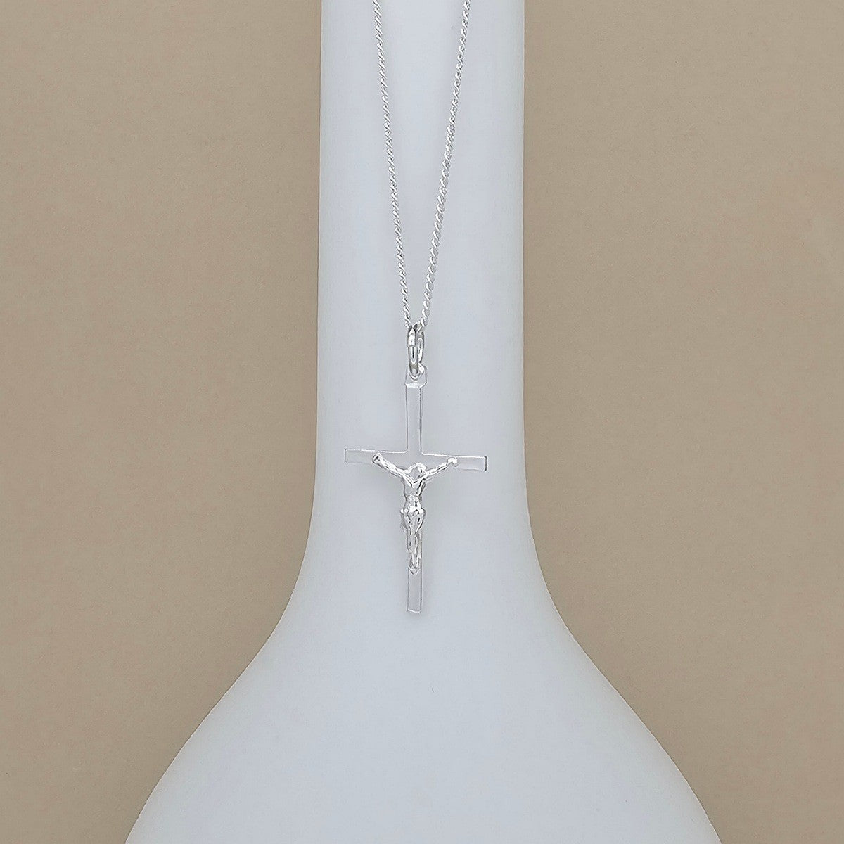A silver crucifix pendant necklace displayed on a white ceramic vase against a solid beige background.