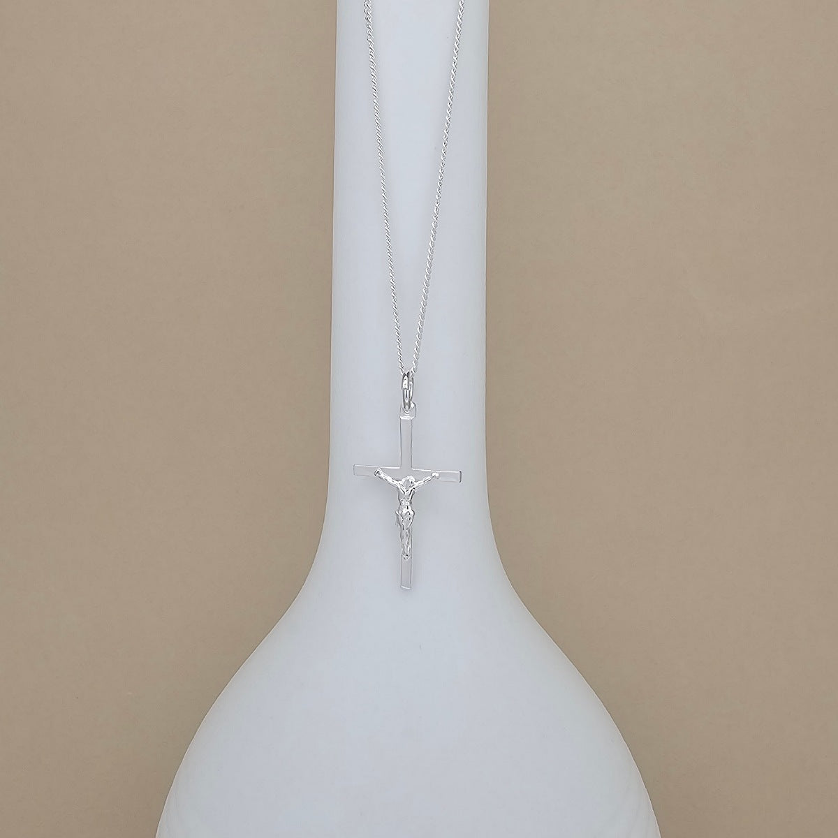 A silver crucifix pendant necklace displayed on a white ceramic vase against a solid beige background.