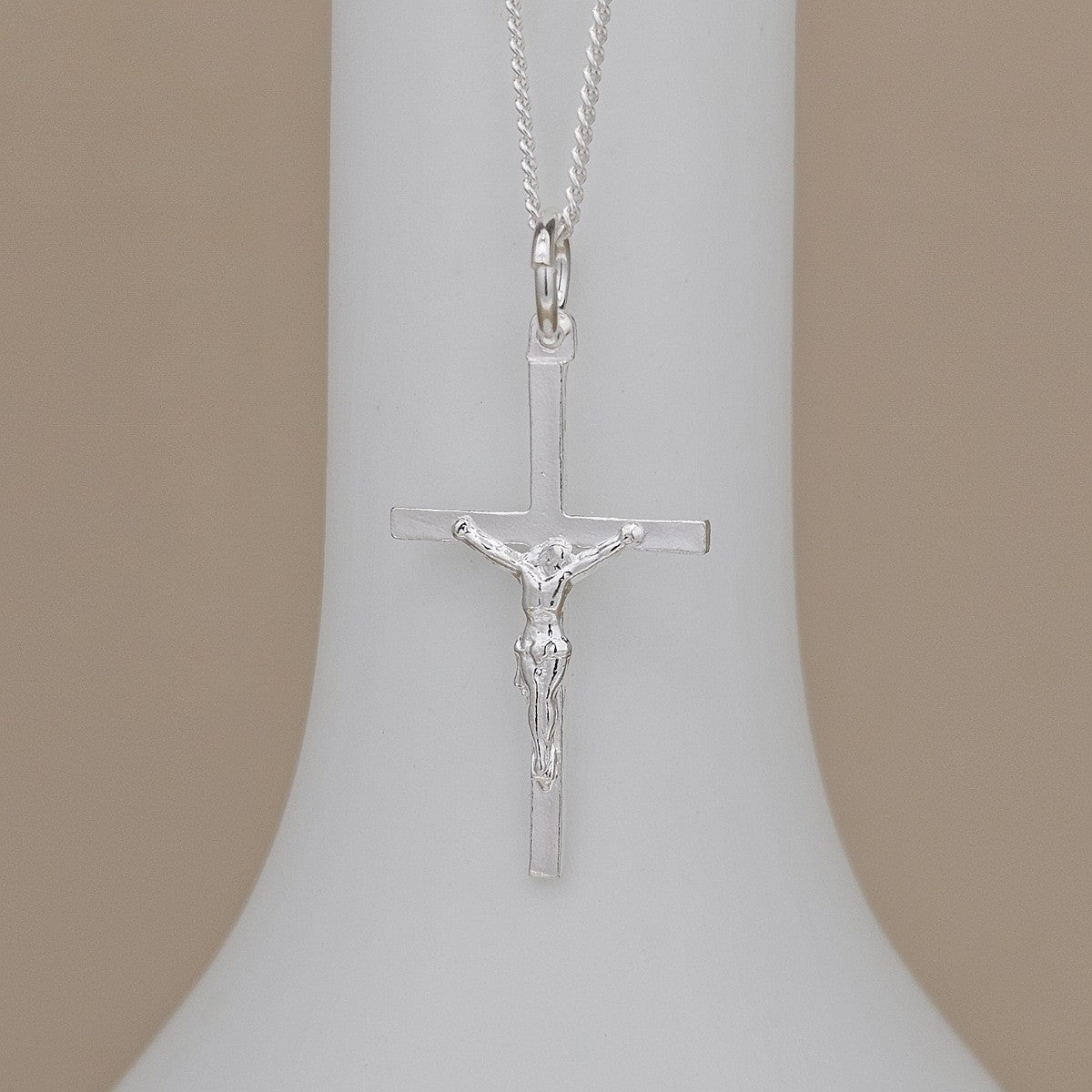 A silver crucifix pendant necklace with a detailed figure of Jesus on a plain cross, hanging from a delicate silver chain against a white display bust with a beige background.