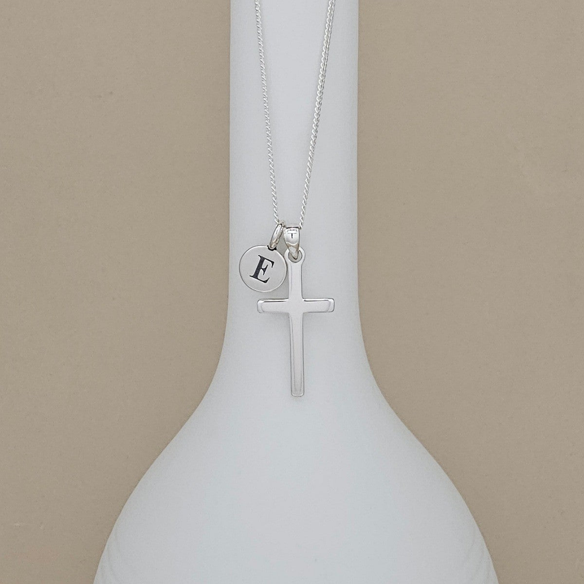A silver cross necklace with a small circular initial pendant engraved with the letter 'E', displayed on a white ceramic vase against a beige background.