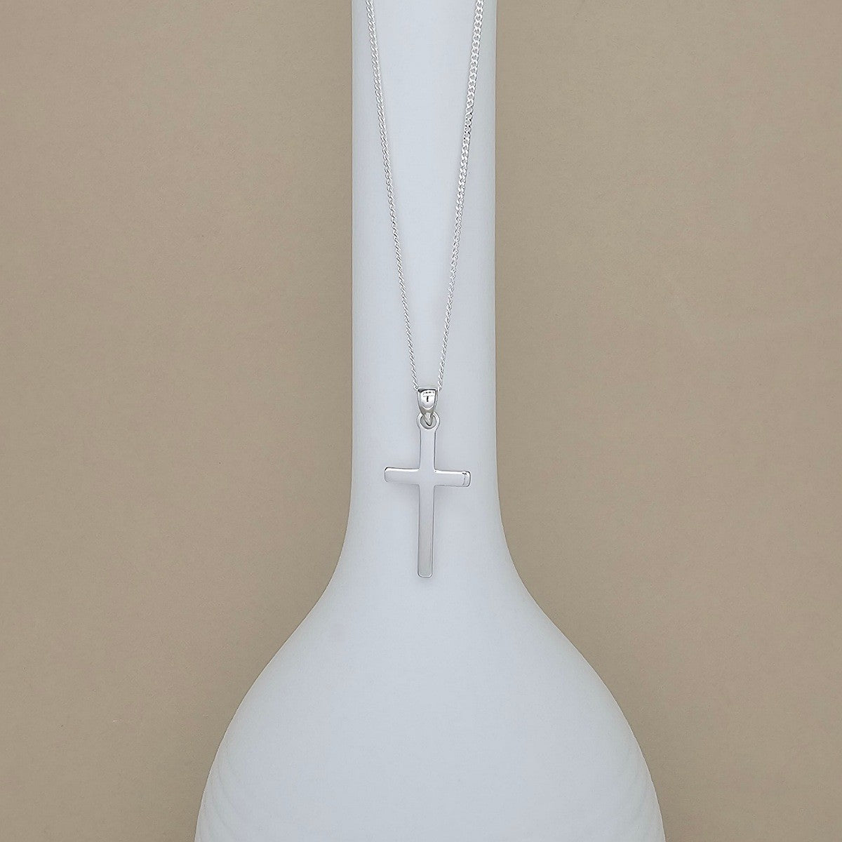 A silver cross pendant necklace hanging from a delicate silver chain, displayed on a white ceramic vase against a solid beige background.
