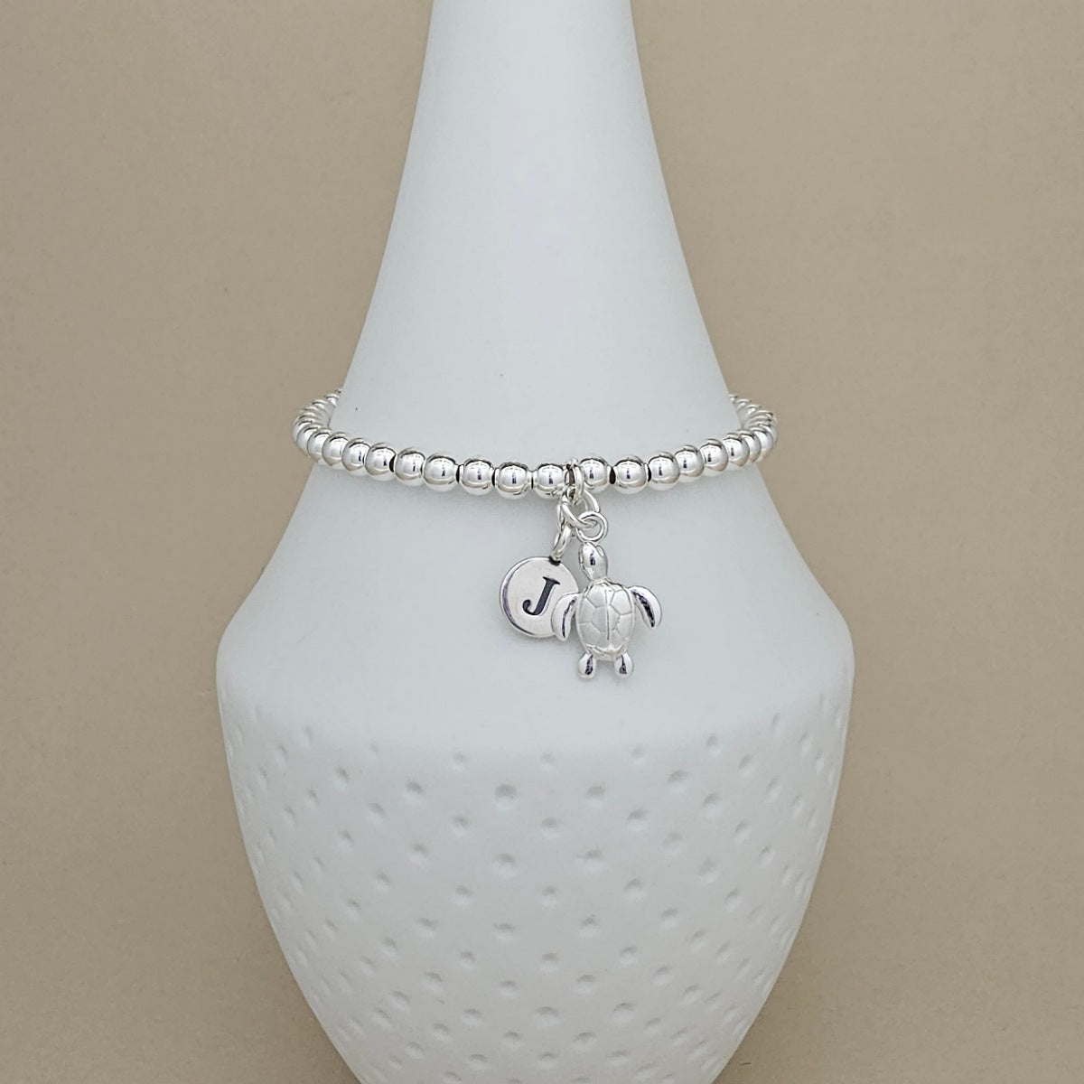 A silver beaded bracelet with a sea turtle charm and a letter 'J' initial charm, displayed on a white textured cone stand against a beige background.