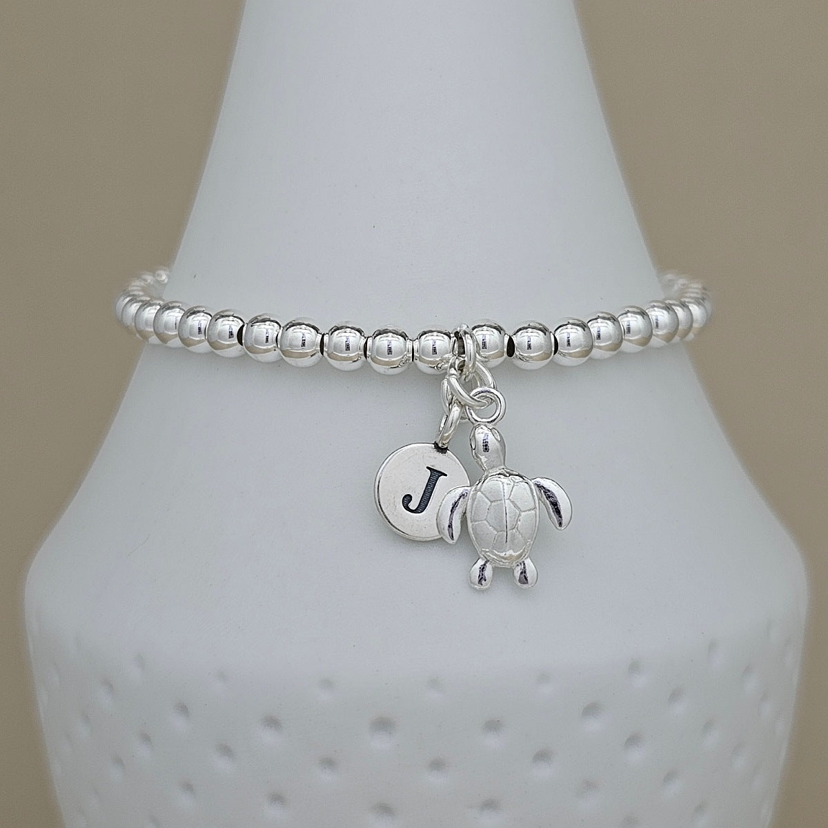 A silver beaded stretch bracelet with a turtle charm and a round initial 'J' monogram charm, displayed on a white textured cone stand.