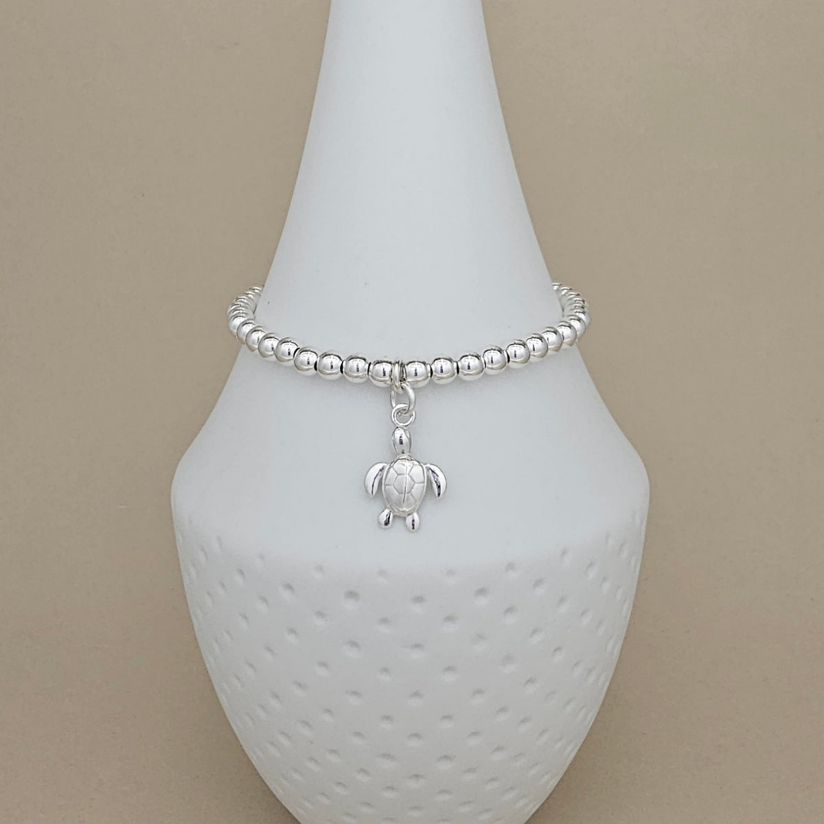 A silver beaded bracelet with a small sea turtle charm, displayed on a white ceramic cone stand against a solid beige background.