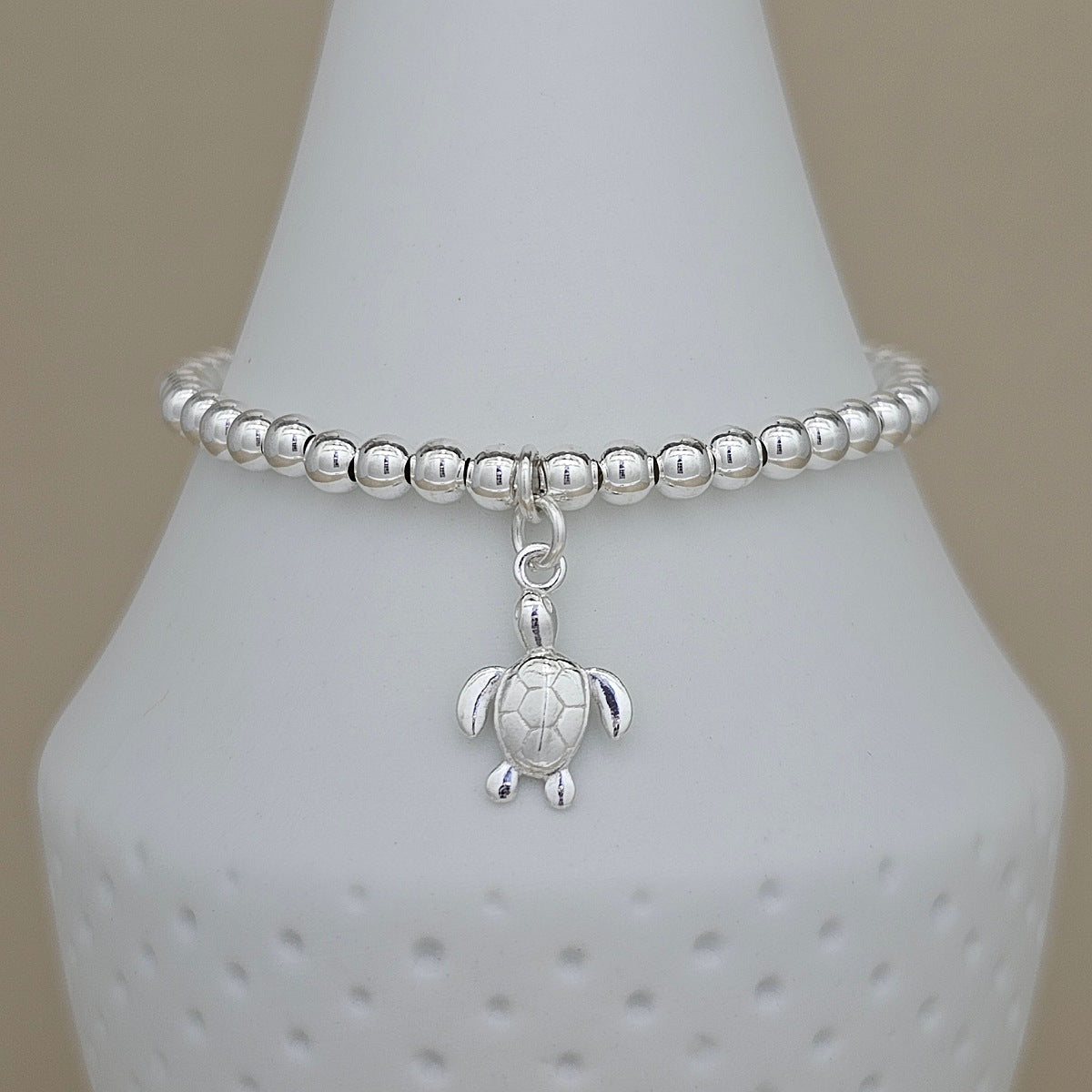 A silver beaded bracelet with a small sea turtle charm, displayed on a white ceramic cone-shaped stand against a neutral beige background.