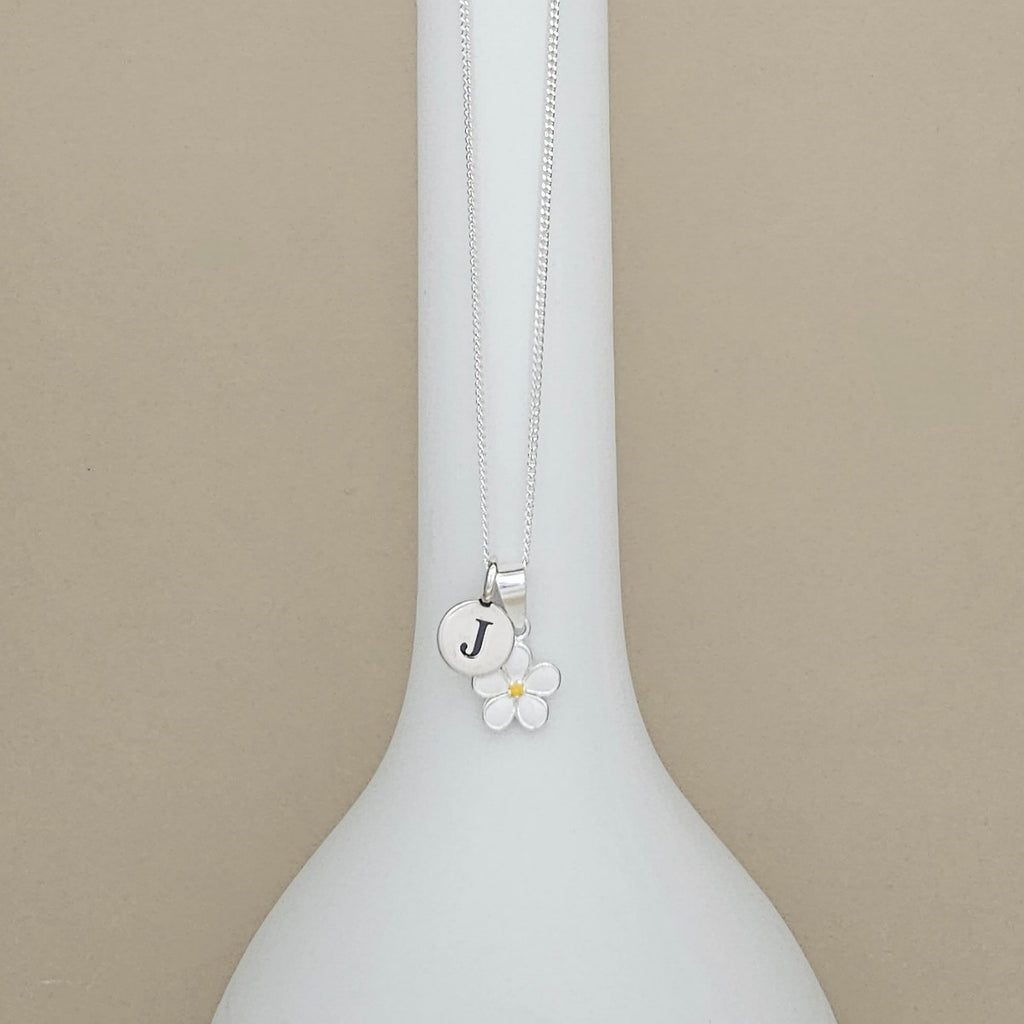 A silver necklace with a round initial 'J' disc charm and a small white flower charm, displayed on a white vase against a beige background.
