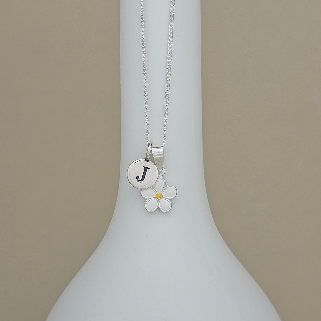 A silver necklace with a round initial 'J' charm and a white flower charm with a yellow center, displayed on a white vase against a beige background.