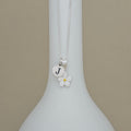 A silver necklace with a round initial 'J' charm and a white flower charm with a yellow center, displayed on a white vase against a beige background.